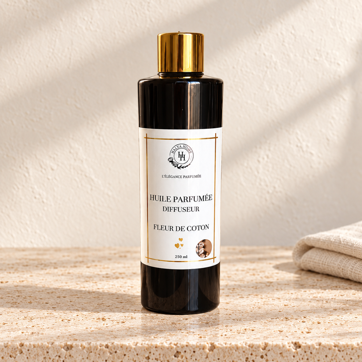 Huile parfumée Fleur de Coton Halya Home pour diffuseur électrique, flacon noir de 250 ml à bouchon doré sur du travertin clair dans un décor ivoire.