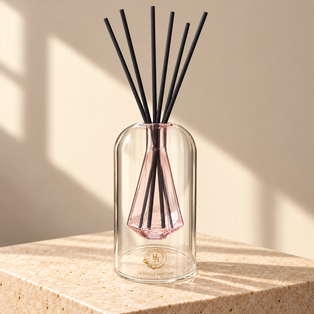 Diffuseur à roseaux Sculpt 02 HALYA HOME de 110 ml, en verre transparent avec réservoir facetté rose et six bâtonnets noirs, sur du travertin.