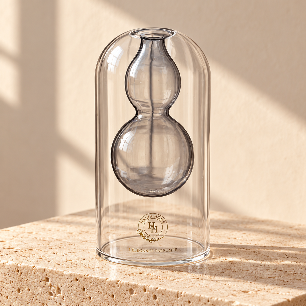 Flacon Sculpt 04 HALYA HOME de 145 ml, enveloppe transparente et réservoir suspendu en verre gris fumé à deux bulbes, sur du travertin clair.