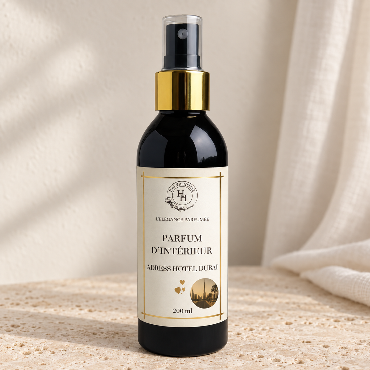 Spray d’intérieur Adress Dubaï Halya Home 200 ml, flacon noir à bague dorée et capuchon transparent sur une pierre claire dans un décor ivoire.