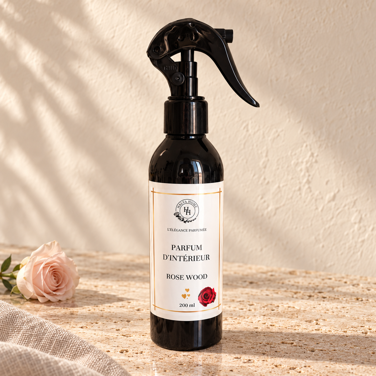 Spray d’intérieur Rose Wood Halya Home 200 ml avec pulvérisateur noir, posé sur une pierre claire près d’une rose.