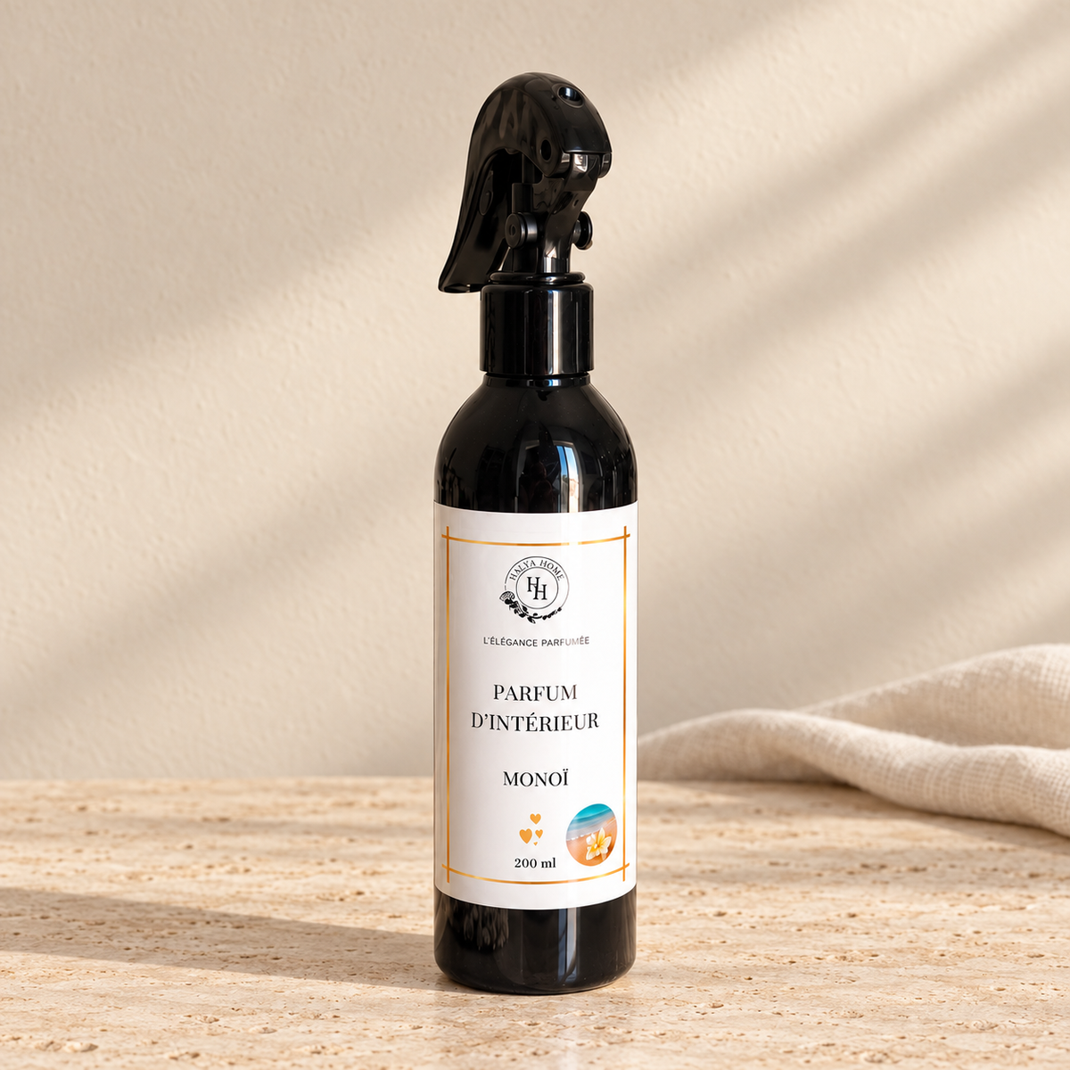 Spray d’intérieur Monoï Halya Home 200 ml, flacon noir avec gâchette sur du travertin clair dans un décor ivoire.