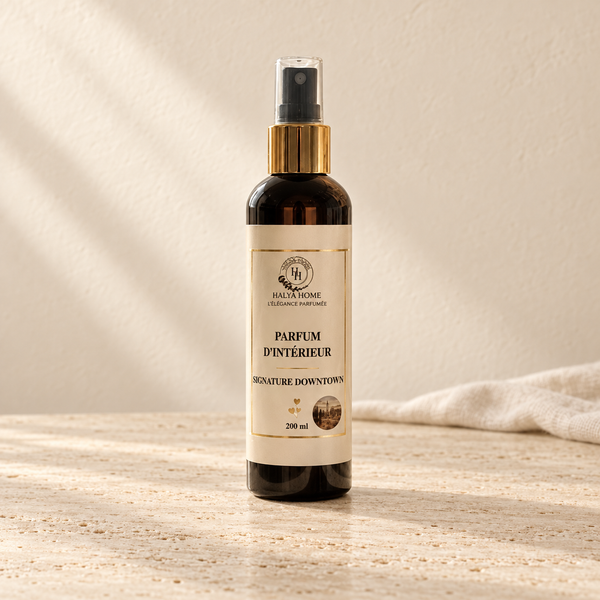 Spray d’intérieur Signature Downtown Halya Home 200 ml, flacon sombre à bague dorée et capuchon transparent sur du travertin dans un décor ivoire.