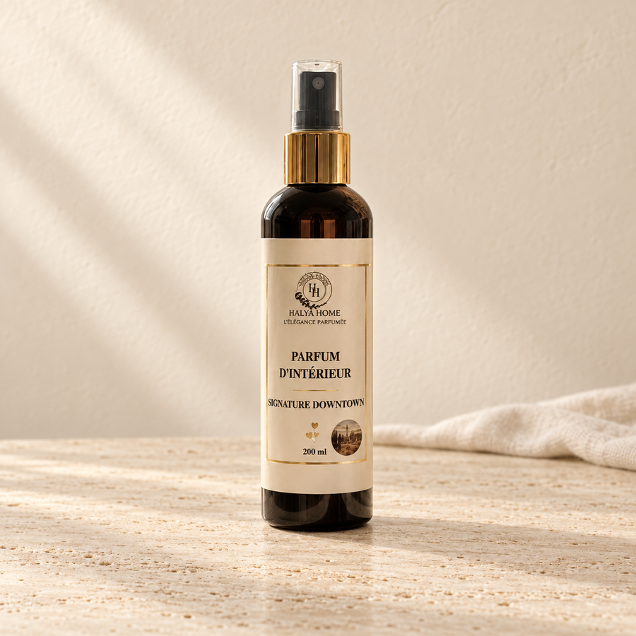 Spray d’intérieur Signature Downtown Halya Home 200 ml, flacon sombre à bague dorée et capuchon transparent sur du travertin dans un décor ivoire.